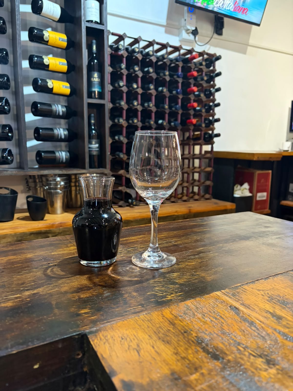 A glass and carafe of red wine on the bar in front of the wine racks at Gavin's Trattoria